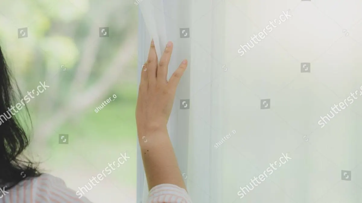 A hand drawing back a sheer white curtain, soft green daylight beyond